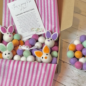 May include: A box filled with colorful felt balls and bunny shapes, ready to be assembled into a garland. The box is lined with pink and white striped paper. The instructions for making the garland are included on a separate card. This is a craft kit for making a felt ball garland.