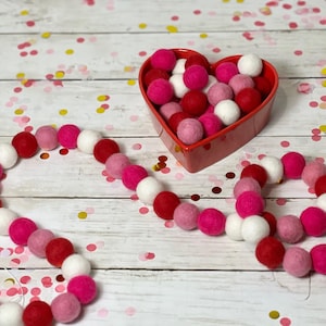 Könnte beinhalten: Eine Girlande aus rosa, roten und weißen Filzkugeln, die aneinandergereiht sind. Die Girlande liegt auf einer weißen Holzoberfläche mit rosa und goldenen Konfetti. Im Hintergrund befindet sich eine rote herzförmige Schale, gefüllt mit weiteren rosa, roten und weißen Filzkugeln.