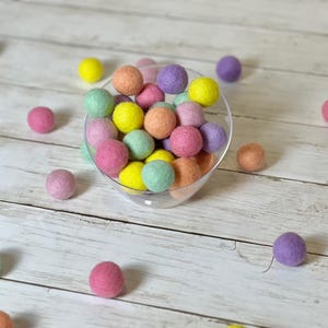 May include: A bowl filled with pastel colored felt balls in shades of pink, yellow, green, and purple. The bowl is sitting on a white wooden surface.