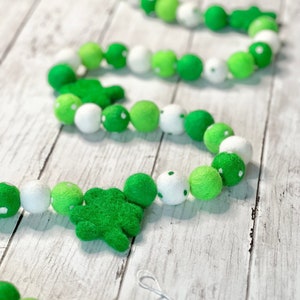 Op de afbeelding: Een groene en witte viltballenkrans met klavertjevier-uitsnijdingen. De krans is geregen aan een wit touwtje en is perfect voor St. Patrick's Day decoratie.