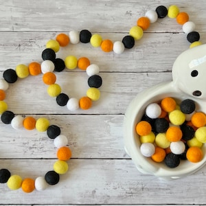 May include: A Halloween garland made of felt balls in orange, yellow, black, and white colors. The garland is arranged in a circle shape and is displayed on a white wooden surface. A white ceramic ghost bowl filled with felt balls in the same colors is also shown.