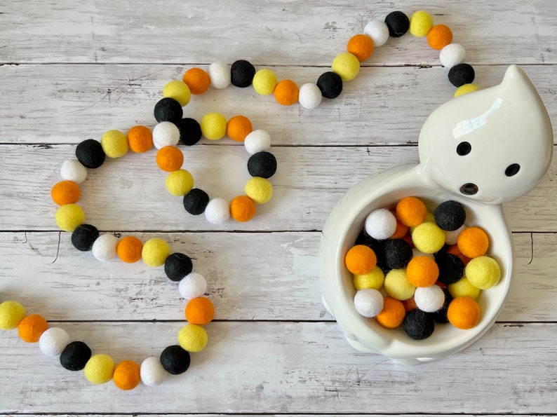 May include: A Halloween garland made of felt balls in orange, yellow, black, and white colors. The garland is arranged in a circle shape and is displayed on a white wooden surface. A white ceramic ghost bowl filled with felt balls in the same colors is also shown.