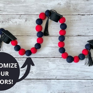 Può includere: Una ghirlanda di laurea realizzata con palline di feltro rosso e blu scuro con cappelli di laurea neri e nappe. La ghirlanda è esposta su una superficie di legno bianco. "Personalizza i tuoi colori!" è scritto in bianco su un cerchio nero.