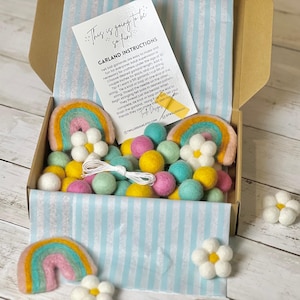 May include: A box containing a DIY felt ball garland kit. The kit includes colorful felt balls in yellow, pink, and teal, felt rainbows, white felt flowers, and instructions. The box has a blue and white striped lining.