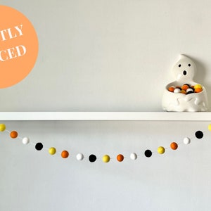 May include: A white shelf with a white ghost-shaped candy dish filled with orange, yellow, and black candy. A garland of orange, yellow, black, and white pom-poms hangs below the shelf. The text "TIGHTLY SPACED" is written on a peach-colored circle.