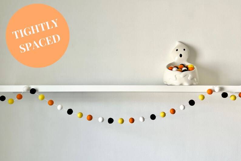 May include: A white shelf with a white ghost-shaped candy dish filled with orange, yellow, and black candy. A garland of orange, yellow, black, and white pom-poms hangs below the shelf. The text "TIGHTLY SPACED" is written on a peach-colored circle.