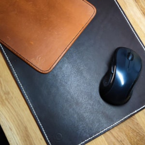Puede incluir: Un mouse pad de cuero marrón con un cuadrado de cuero marrón a juego encima y un ratón inalámbrico negro. La alfombrilla del ratón es negra con costuras blancas en los bordes.