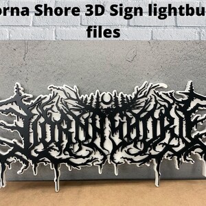 May include: A black and white 3D sign with the text "Lorna Shore" in a stylized font. The sign is made of wood and has a lightburn finish. The sign is mounted on a white wall.