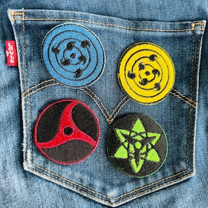May include: Four round embroidered patches with different designs on a blue denim background. The patches are black, red, green, and yellow with black outlines. The patches are all different sizes and are arranged in a square shape.
