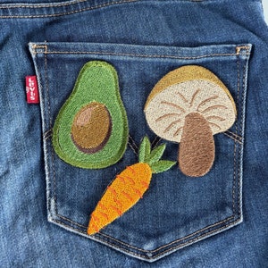 May include: A pair of dark blue denim jeans featuring a back pocket embroidered with three distinct patches: a green avocado with a brown pit, a tan and brown mushroom, and an orange carrot with green leafy tops. A small red Levi's tag is visible.