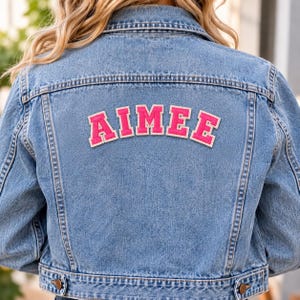 May include: A light blue denim jacket featuring the name AIMEE in bright pink varsity-style letters with white trim across the upper back. The jacket has a classic collar, long sleeves, and visible stitching details along the seams and hemline.