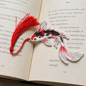 May include: An embroidered bookmark shaped like a koi fish with white, black, and red thread detailing. The fish features a long, vibrant red tassel attached to its head by a matching red cord, resting flat against the open pages of a book.