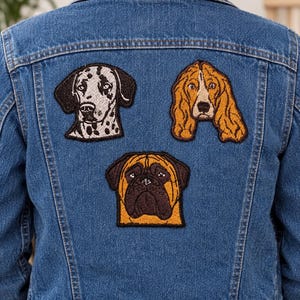 May include: A blue denim jacket featuring three embroidered dog patches on the back. The patches depict a white and black spotted Dalmatian, a golden-brown spaniel, and a tan and dark brown mastiff, all arranged in a triangular formation.