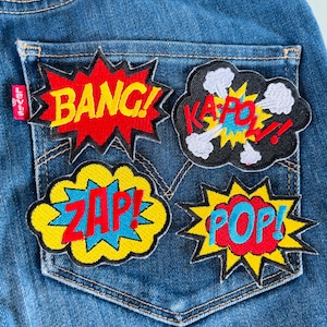May include: Four colourful embroidered patches with comic book sound effects: "Bang!" in red, "Zap!" in yellow, "Pop!" in red, and "Kapow!" in black. The patches are sewn onto a blue denim pocket.