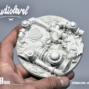 May include: A detailed, circular miniature base with industrial elements, including pipes, a manhole cover, and debris. The base is 100mm in diameter and features the text "studiolevel" and "handmade for wargames fanatics."