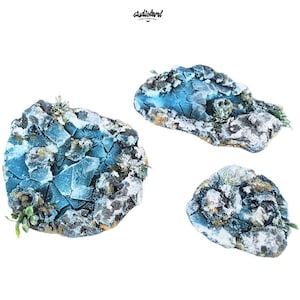 May include: Three miniature terrain pieces featuring cracked blue ice surfaces surrounded by rocky, snow-dusted edges. Each piece displays textured stone formations with white highlights and small patches of green vegetation, set against a plain white background.