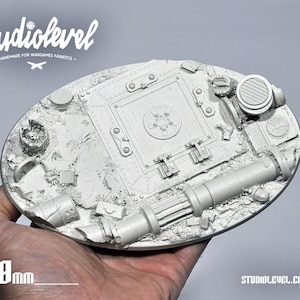 Könnte beinhalten: Eine detaillierte, graue Miniatur-War-Game-Basis, 17,8 cm lang. Die Basis weist komplizierte mechanische und industrielle Elemente auf, darunter eine zentrale Luke, Rohre und strukturiertes Gelände. Der Text "studiolevel" ist sichtbar.