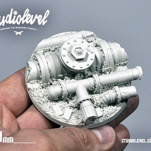 May include: A 68 mm diameter round, gray resin base with a detailed, textured design of pipes and valves. The base is designed for use in tabletop wargaming.