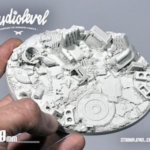 May include: A 120mm diameter round, gray, resin base for miniature wargaming. The base features a detailed, textured surface with a variety of debris and rubble, including a chair, a trash can, and a broken window. The base is designed to create a realistic and immersive battlefield environment.