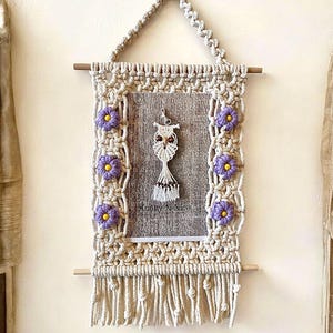 May include: A macrame wall hanging with a rectangular frame, adorned with purple and yellow flower accents. The frame surrounds a wooden panel with a macrame owl pendant. The piece is suspended by a woven triangle and a wooden dowel.