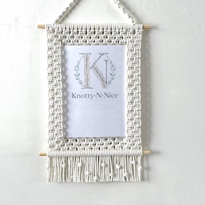 May include: A macrame wall hanging with a rectangular frame, wooden dowels, and a fringe. The frame holds a white print with a floral "K" design and the text "Knotty-N-Nice". The macrame is off-white and hangs from a knotted loop.