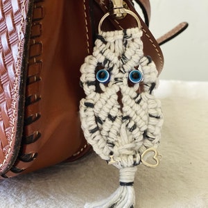 May include: A macrame owl keychain with blue glass eyes and a silver-colored clasp. The owl is white with black flecks and a small tassel. A silver heart charm hangs from the bottom. The keychain is attached to a brown leather bag.