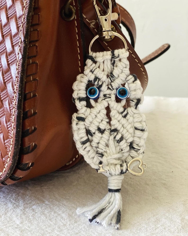 May include: A macrame owl keychain with blue glass eyes and a silver-colored clasp. The owl is white with black flecks and a small tassel. A silver heart charm hangs from the bottom. The keychain is attached to a brown leather bag.