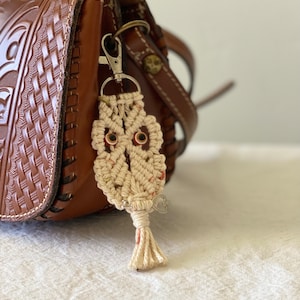 May include: A handmade macrame owl keychain with brown eyes and a small silver heart charm. The owl is attached to a silver clasp, hanging next to a brown leather purse with a woven design.