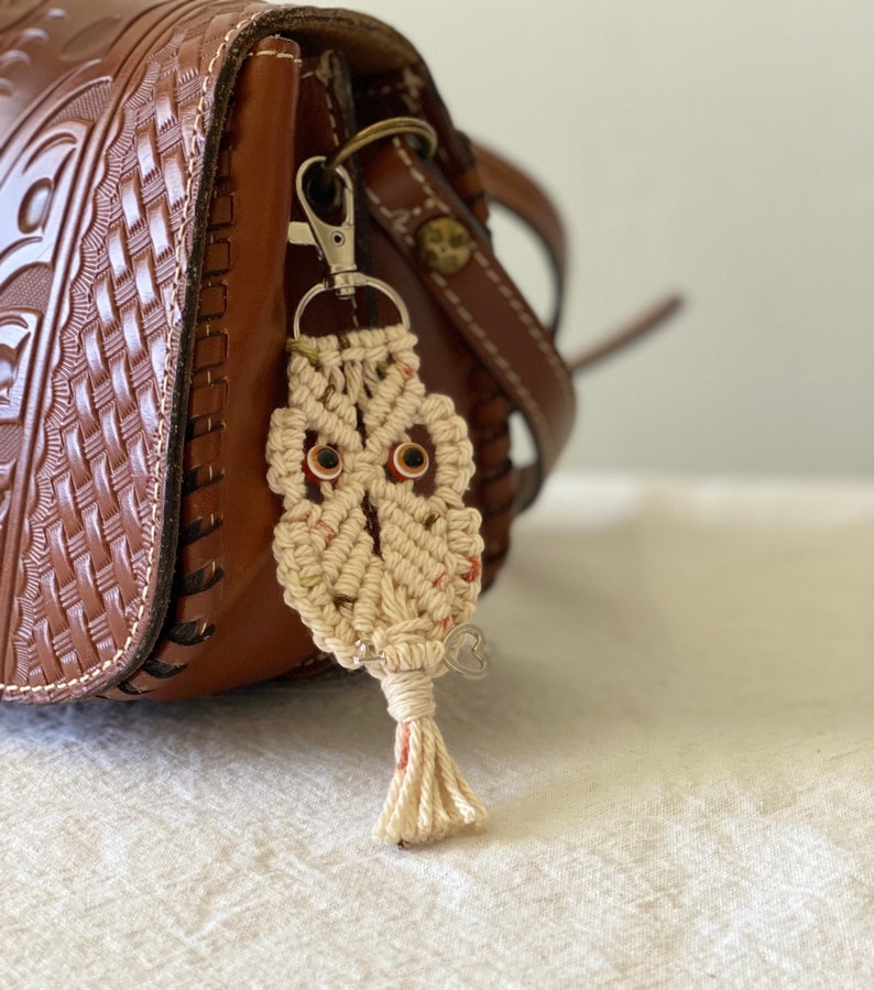 May include: A handmade macrame owl keychain with brown eyes and a small silver heart charm. The owl is attached to a silver clasp, hanging next to a brown leather purse with a woven design.