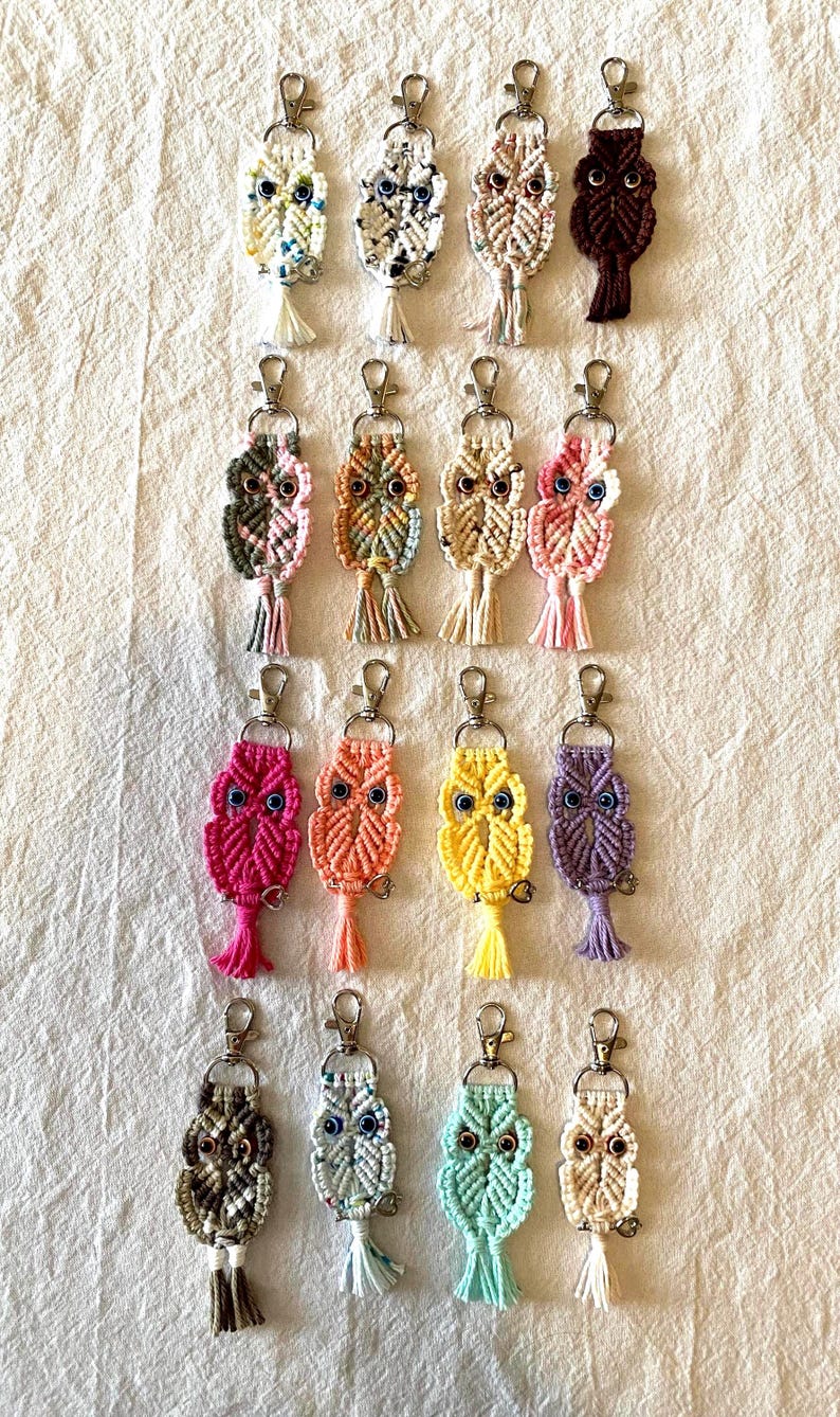 May include: A collection of macrame owl keychains in various colors, including pink, orange, yellow, and purple. Each keychain features a silver clasp and a tassel. The owls have detailed eyes and are arranged on a white fabric background.