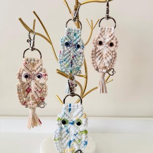 May include: Four macrame owl keychains in various colors, including white, pink, and blue, hang from a gold-colored decorative stand. Each owl has large, googly eyes and a tassel. The keychains are a charming, handmade accessory.
