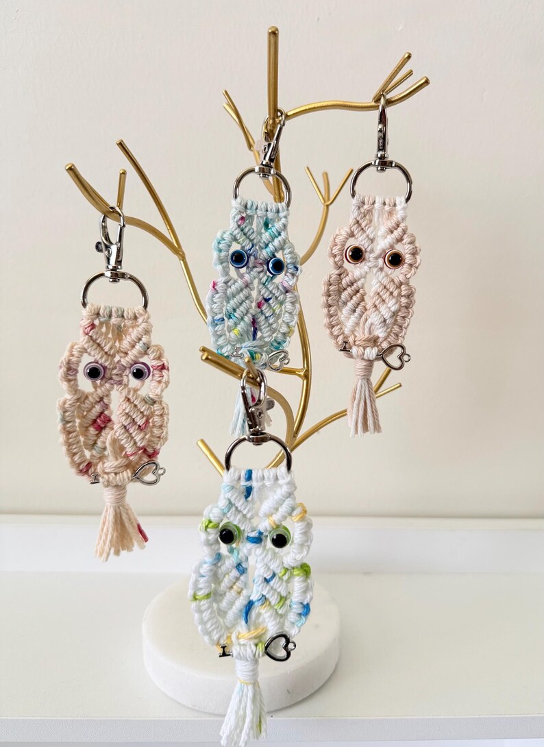 May include: Four macrame owl keychains in various colors, including white, pink, and blue, hang from a gold-colored decorative stand. Each owl has large, googly eyes and a tassel. The keychains are a charming, handmade accessory.