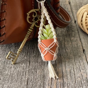 May include: A small potted plant with green leaves hangs from a white macrame keychain attached to a brown leather bag.