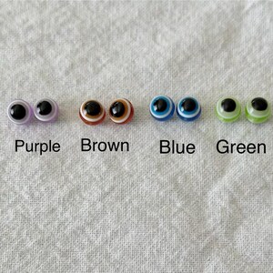 May include: A collection of four pairs of glass eye cabochons in purple, brown, blue, and green. Each pair has a black pupil and a colored iris, with the color names printed below each pair.