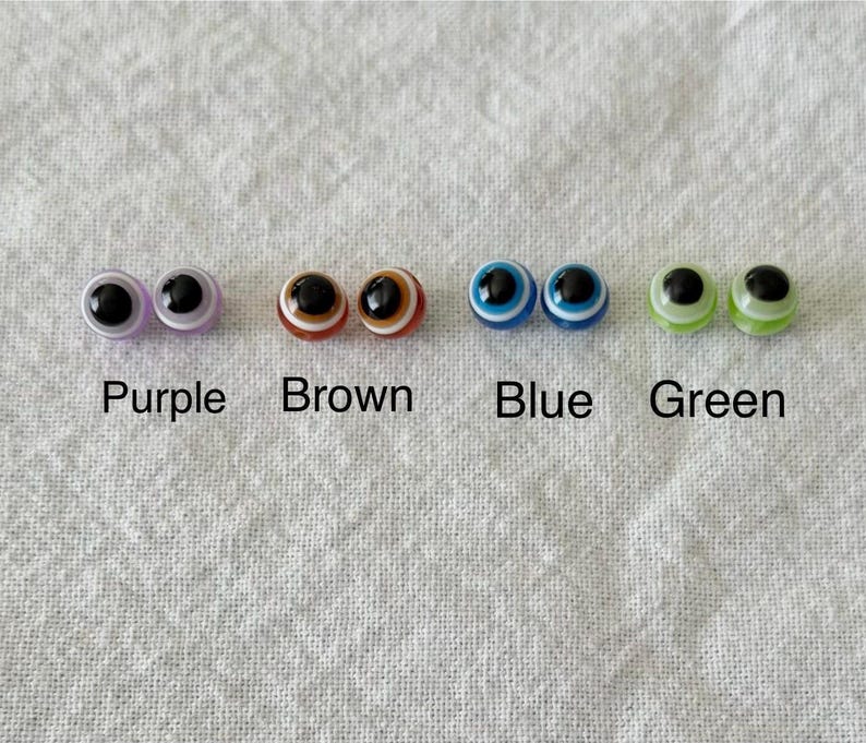May include: A collection of four pairs of glass eye cabochons in purple, brown, blue, and green. Each pair has a black pupil and a colored iris, with the color names printed below each pair.