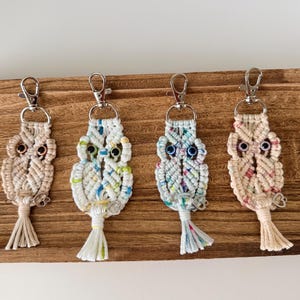 May include: Four macrame owl keychains in beige, white, and blue hues, each with a silver clasp and tassel. The owls have detailed eyes and are displayed on a wooden surface, showcasing a handcrafted, bohemian style.