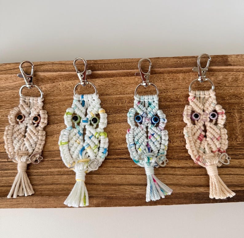 May include: Four macrame owl keychains in beige, white, and blue hues, each with a silver clasp and tassel. The owls have detailed eyes and are displayed on a wooden surface, showcasing a handcrafted, bohemian style.