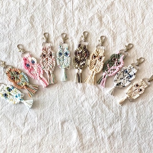 May include: A collection of handmade macrame owl keychains in various colors. Each keychain features an owl design with detailed knotting, a metal clasp, and a tassel. The color palette includes pink, blue, green, and neutral tones.