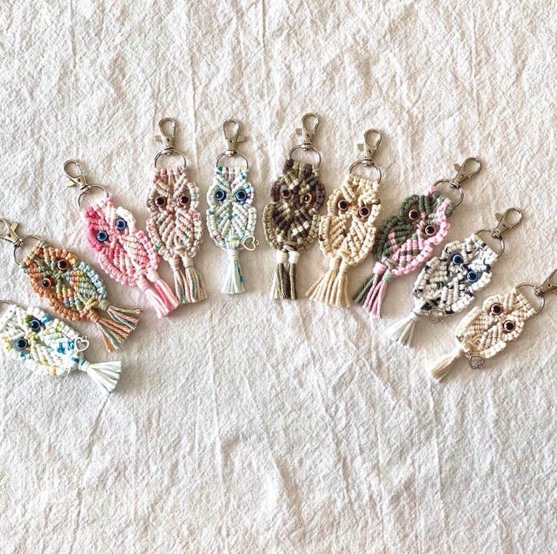 May include: A collection of handmade macrame owl keychains in various colors. Each keychain features an owl design with detailed knotting, a metal clasp, and a tassel. The color palette includes pink, blue, green, and neutral tones.