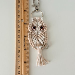 May include: A handmade macrame owl keychain in cream and tan colors. The owl has brown eyes and a small silver key charm. The keychain is approximately 3 inches long, excluding the metal clasp and tassel.