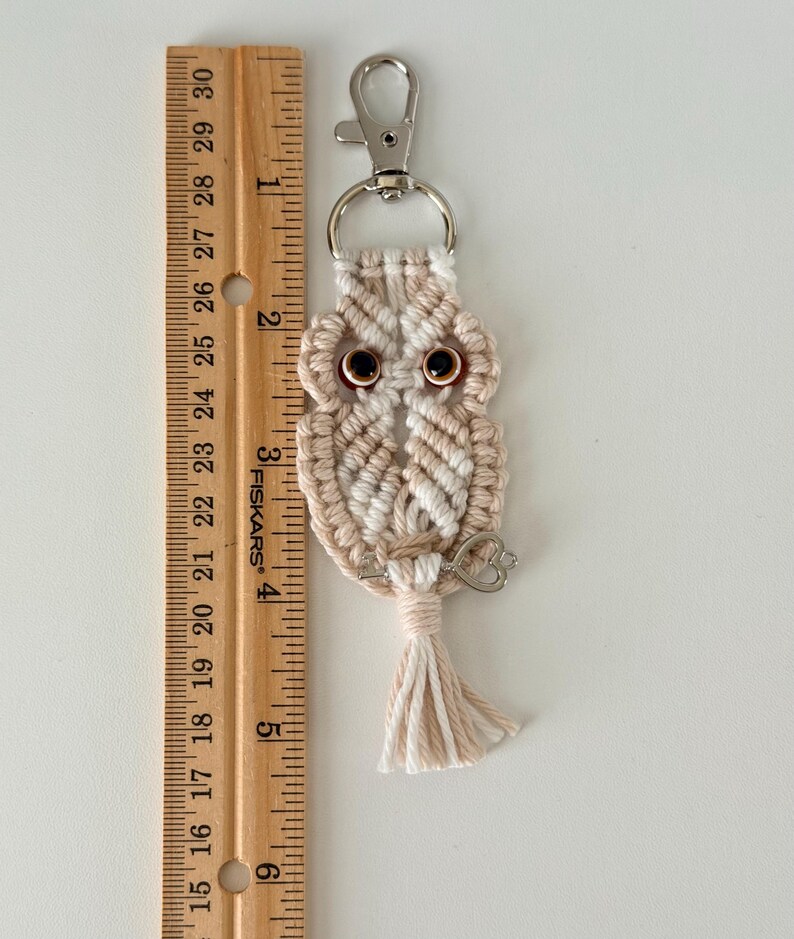 May include: A handmade macrame owl keychain in cream and tan colors. The owl has brown eyes and a small silver key charm. The keychain is approximately 3 inches long, excluding the metal clasp and tassel.