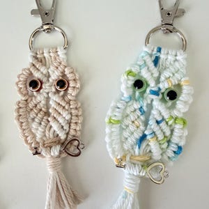 May include: Two macrame owl keychains. One is beige and white, the other is white with blue and green accents. Each has black eyes, a silver clasp, and a heart charm. The owl keychains are handmade.