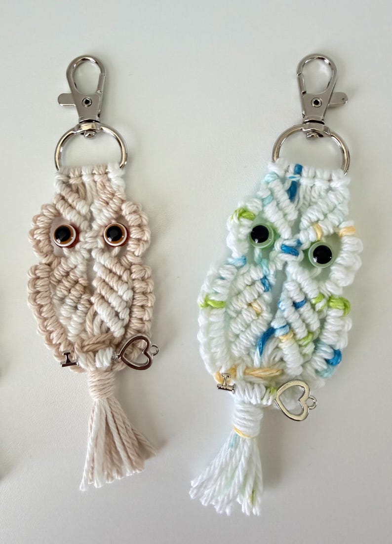 May include: Two macrame owl keychains. One is beige and white, the other is white with blue and green accents. Each has black eyes, a silver clasp, and a heart charm. The owl keychains are handmade.