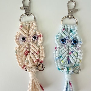 May include: Two macrame owl keychains. One is cream-colored with pink and red accents, the other is light blue with colorful flecks. Both have silver-tone clasps, googly eyes, and heart charms.