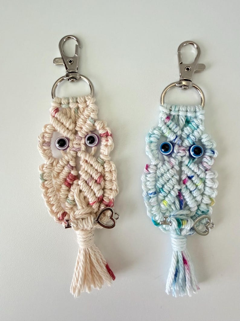 May include: Two macrame owl keychains. One is cream-colored with pink and red accents, the other is light blue with colorful flecks. Both have silver-tone clasps, googly eyes, and heart charms.