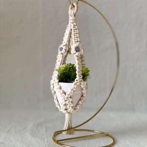 May include: A cream-colored macrame plant hanger with a small white pot holding a green succulent. The hanger is adorned with purple beads and suspended from a gold-colored metal stand. The stand has a curved design.
