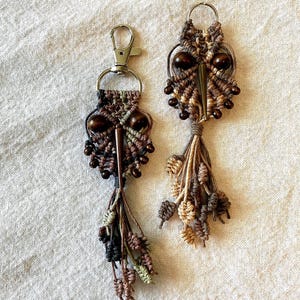 May include: Two owl-shaped keychains with brown, beige, and green tassels. The owls are made of macrame and have wooden beads for eyes.