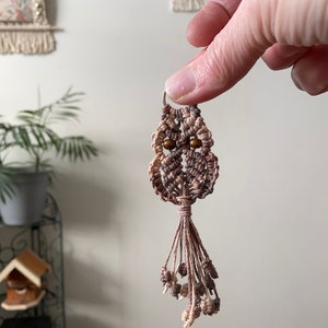 May include: A small macrame owl keychain with a brown and pink color gradient. The owl has brown wooden bead eyes and a long tassel.