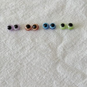 May include: A collection of six pairs of plastic craft eyes in various colors. The eyes have black pupils and colored irises, including purple, brown, blue, and green. They are arranged in a row against a white fabric background.