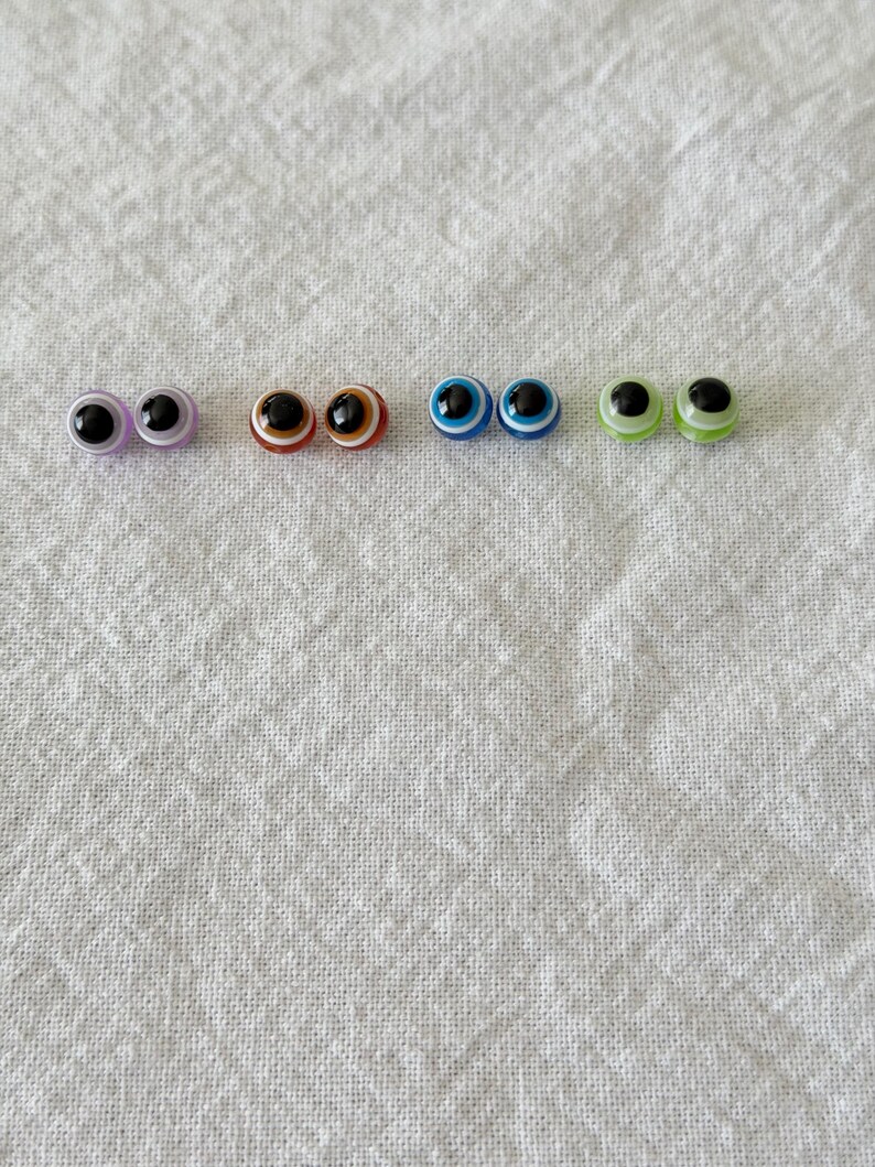May include: A collection of six pairs of plastic craft eyes in various colors. The eyes have black pupils and colored irises, including purple, brown, blue, and green. They are arranged in a row against a white fabric background.